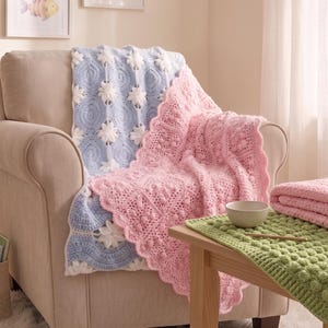 May include: A light blue and white crocheted blanket with a floral pattern draped over a chair, alongside a pink crocheted blanket with a scalloped edge and a green textured crocheted mat featuring a bobble stitch pattern on a wooden table.