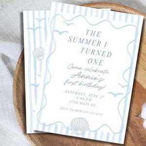 May include: A rectangular birthday invitation card featuring light blue vertical stripes, a wavy border, and illustrations of seashells and seagulls. The text reads: THE SUMMER I TURNED ONE, Come celebrate Annie's first birthday, SATURDAY, JUNE 27, 3:00 PM, 1234 MAIN ST., RSVP TO MOM 555-123-4567.