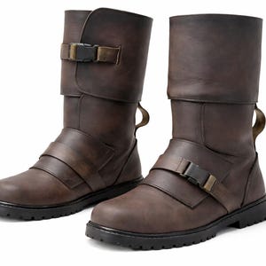 May include: A pair of brown leather boots with a rugged design. The boots feature multiple straps with metal buckles and a folded-over top. The soles are black and the boots appear to be new.