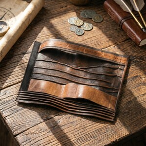 May include: A handcrafted leather journal with a unique, layered design. The journal features a combination of dark brown and tan leather, with a textured, wavy pattern. The cover is folded and secured with a wide leather strap. The journal is on a wooden surface.