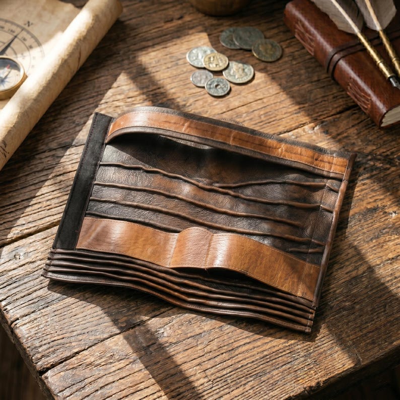 May include: A handcrafted leather journal with a unique, layered design. The journal features a combination of dark brown and tan leather, with a textured, wavy pattern. The cover is folded and secured with a wide leather strap. The journal is on a wooden surface.