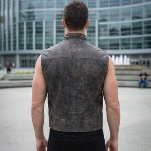 Op de afbeelding: Een mouwloos vest van verweerd donkerbruin leer met een hoge kraag, zichtbare naaddetails op de rug en een rechte zoom. Het kledingstuk wordt gedragen over een zwarte broek en heeft een getextureerde afwerking en een gestructureerde pasvorm.