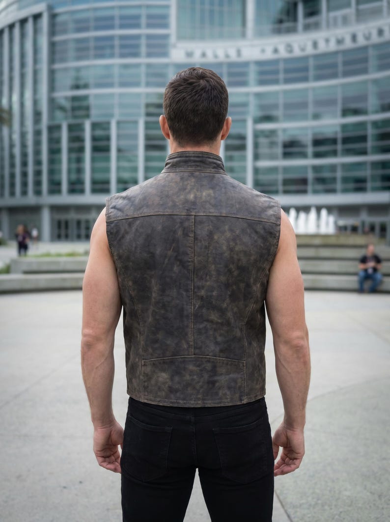 Op de afbeelding: Een mouwloos vest van verweerd donkerbruin leer met een hoge kraag, zichtbare naaddetails op de rug en een rechte zoom. Het kledingstuk wordt gedragen over een zwarte broek en heeft een getextureerde afwerking en een gestructureerde pasvorm.