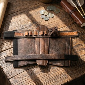 May include: A distressed brown leather arm guard with multiple small pouches and a small bag. The arm guard has a wide strap with black velcro closures. The leather has a worn, vintage appearance, suggesting a historical or fantasy theme.