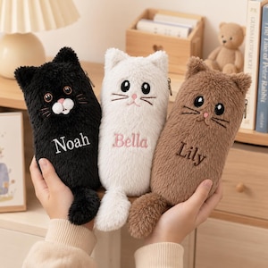 Peut inclure: Trois trousses à crayons en peluche en forme de chat, de couleurs noire, blanche et marron. Chaque trousse présente des détails faciaux brodés, une petite queue et un prénom personnalisé cousu sur le devant : Noah, Bella et Lily.