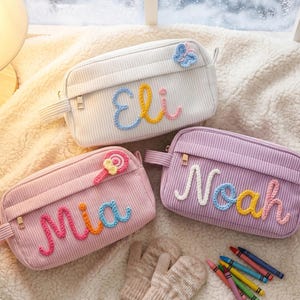 May include: Three corduroy pencil cases in white, pink, and lavender, each with a name embroidered in colorful yarn. The white case has a butterfly, the pink one a lollipop, and the lavender one has no embellishment. A pair of mittens and crayons are also visible.
