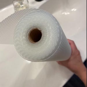 May include: A roll of clear, textured shelf liner. The liner is made of a woven material and is rolled up, revealing a brown cardboard core. The liner is held in front of a white sink.