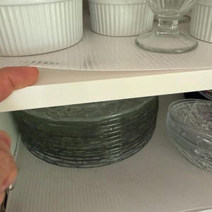 May include: A cabinet shelf filled with white ceramic ramekins, clear glass dessert dishes, and a stack of green-tinted glass plates. The shelf is lined with a light-colored, textured shelf liner.