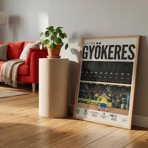 Puede incluir: Un póster enmarcado con el nombre "GYOKERES" en letras negras grandes. El póster incluye una imagen de un jugador de fútbol con una camiseta amarilla, con el texto "VICTOR" encima del nombre. Una pequeña planta en maceta está sobre una mesa auxiliar cilíndrica.