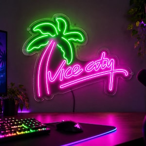 Könnte beinhalten: Ein an der Wand montiertes Neonschild mit einer leuchtend grünen Palme und dem Schriftzug Vice City in einer kräftigen pinkfarbenen Schreibschrift. Das Schild ist auf einer transparenten Acrylplatte befestigt und leuchtet in einem dunklen Raum.