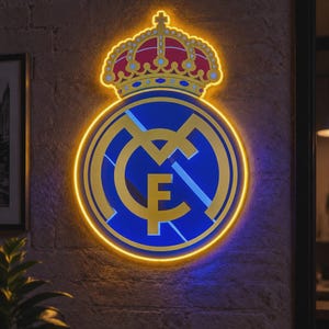 以下が含まれることがあります： レアル・マドリードのサッカークラブのロゴをあしらった円形のネオンサインで、明るい黄色の輪郭で縁取られています。デザインには、MCFの文字が絡み合った金色の紋章が含まれており、その上部には赤、青、金の詳細な王冠が飾られています。