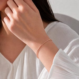Può includere: Un braccialetto sottile color oro rosa con un delicato design a catena testurizzata. Il braccialetto presenta un profilo circolare liscio che poggia comodamente attorno al polso, mostrando una finitura metallica che cattura la luce.