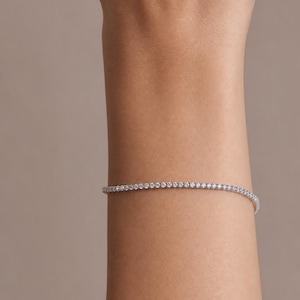 Puede incluir: Una delicada pulsera de tenis que presenta una hilera continua de pequeñas piedras redondas, claras y brillantes, engastadas en una fina banda de metal plateado que rodea la muñeca, creando un aspecto refinado y elegante con cierre.