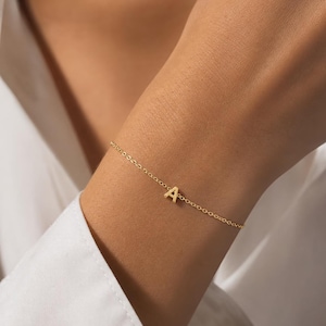 Puede incluir: Una delicada pulsera de cadena en tono dorado que presenta un dije central con la letra A mayúscula. La pulsera está construida con eslabones circulares finos y tiene un acabado metálico pulido, creando una pieza de joyería sencilla.