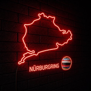 Peut inclure: Une enseigne néon rouge représentant le tracé du circuit du Nürburgring avec le texte NÜRBURGRING en lettres néon rouges et un petit graphique de casque de course détaillé situé à droite du texte sur un mur de briques sombres.