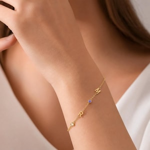 Puede incluir: Una pulsera de cadena en tono dorado con dos letras mayúsculas, M y R, intercaladas con dos pequeñas piedras circulares, una transparente y otra morada. Los delicados eslabones metálicos conectan cada elemento en una línea sobre la muñeca.
