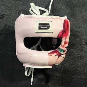 Pode incluir: Capacete de boxe rosa com abertura preta para os olhos e cadarços brancos. O capacete apresenta um design gráfico na lateral e o texto "Creation BY ARSPAINTL". Um logotipo branco e preto está na frente.