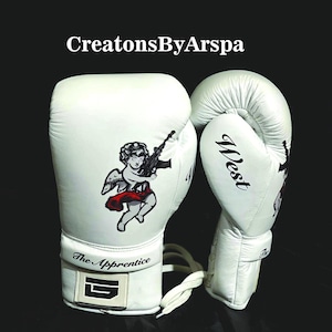 Puede incluir: Guantes de boxeo blancos con un ángel negro y rojo sosteniendo un arma. El texto "The Apprentice" está impreso en la correa de la muñeca. La palabra "West" está impresa en el otro guante.