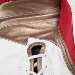 Op de afbeelding: Een bokshandschoen met een combinatie van witte, metallic gouden en rode panelen. De handschoen is voorzien van een vetersluiting met witte veters door metallic gouden oogjes, met zichtbare stiksels langs de naden van de verschillende panelen.