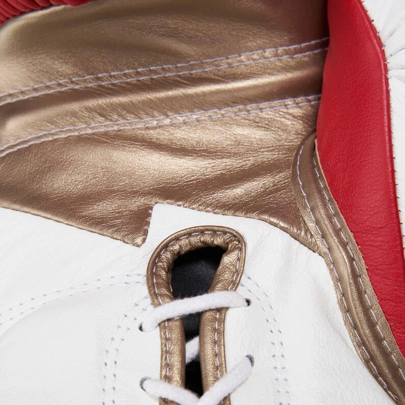 Op de afbeelding: Een bokshandschoen met een combinatie van witte, metallic gouden en rode panelen. De handschoen is voorzien van een vetersluiting met witte veters door metallic gouden oogjes, met zichtbare stiksels langs de naden van de verschillende panelen.