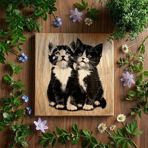 May include: A wooden square featuring a black and white illustration of two kittens. The kittens have white bibs and are set against a light wood grain background. The image is surrounded by greenery, small white daisies, and purple flowers.