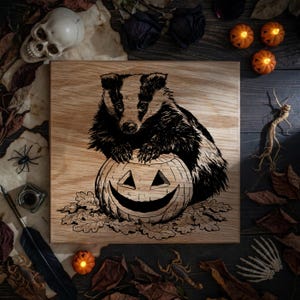 May include: A square wooden plaque featuring a black ink illustration of a badger resting its paws on a carved jack-o'-lantern pumpkin, surrounded by etched oak leaves, presented on a light-colored wood grain surface with a natural finish.
