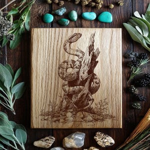 May include: A wooden chopping board featuring a detailed engraving of a snake coiled around a tree branch. The snake is brown and tan, and the wood has a natural grain. Turquoise stones and greenery surround the board.