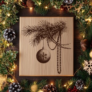 Könnte beinhalten: Eine quadratische Holzplatte mit einer dunkelbraunen, lasergravierten Abbildung eines Tannenzweigs, an dem eine mit einem Schneeflockenmuster verzierte Weihnachtskugel und eine Perlenkette hängen, zentriert auf hellem Holzmaserungshintergrund.