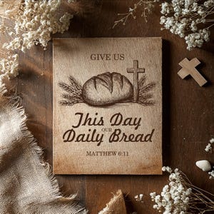 Könnte beinhalten: Holztafel mit dem Text "GIVE US THIS DAY OUR DAILY BREAD" und "MATTHEW 6:11". Die Tafel zeigt eine Brotlaib-, Weizenähren- und Kreuzillustration. Der Hintergrund umfasst Trockenblumen und ein kleines Holzkreuz.
