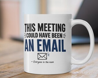 May include: A white ceramic mug featuring the black and blue text THIS MEETING COULD HAVE BEEN AN EMAIL, an envelope icon, and the smaller text - Everyone in the room. The mug has a curved handle on the right side and a smooth, glossy finish.