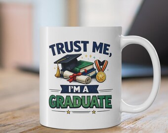 May include: A white ceramic mug featuring a graphic of a black graduation cap, stacked books, a rolled diploma with a red ribbon, and a gold medal. The text reads TRUST ME, I'M A GRADUATE in dark blue and green block lettering with small stars.