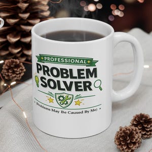 Może przedstawiać: Biały ceramiczny kubek z napisem "PROFESSIONAL PROBLEM SOLVER" pogrubionymi czarnymi literami, któremu towarzyszą zielone ikony żarówki, lupy oraz tarczy z kluczem płaskim i kołem zębatym, a także tekst "Problems May Be Caused By Me".