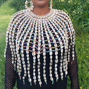 May include: A decorative capelet constructed from numerous off-white cowrie shells arranged in a tiered, web-like pattern. The shells are interconnected by rows of small, multicoloured glass beads, creating a fringe effect that drapes over the shoulders.
