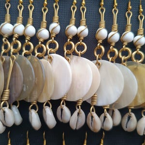 Puede incluir: Primer plano de múltiples pares de pendientes. Cada pendiente presenta un gancho de metal dorado, una cuenta blanca y un colgante de concha circular, plano y grande. Algunos pendientes también incluyen pequeños amuletos de concha blanca.