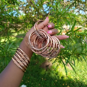 Puede incluir: Una colección de brazaletes de color cobre. Algunos son lisos, mientras que otros presentan intrincados diseños tejidos. Los brazaletes se sostienen en una mano con esmalte de uñas rosa, sobre un fondo de follaje verde y cielo brillante.