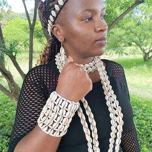 May include: A jewellery set consisting of a cowrie shell headband, a multi-strand cowrie shell necklace, a wide cowrie shell cuff bracelet, and matching cowrie shell drop earrings. Each piece is crafted from white cowrie shells and black thread.