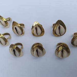 Puede incluir: Un conjunto de ocho anillos, cada uno con una concha de cauri blanca central engastada en un marco de metal dorado en forma de lágrima. Los anillos tienen bandas ajustables y un acabado texturizado y reflectante en el metal.