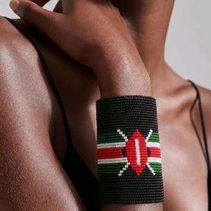 May include: A black beaded cuff bracelet with a red, white, and green design. The bracelet features a central emblem with a red oval and white accents, and horizontal stripes of red, white, and green. The bracelet is worn on a wrist.