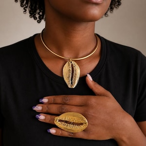 May include: A gold-toned metal choker necklace featuring a central cowrie shell pendant, paired with a matching large gold-toned cowrie shell statement ring. Both pieces exhibit a textured, metallic finish with a distinct central slit design.