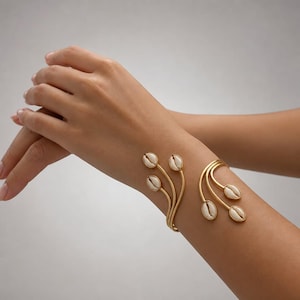 Könnte beinhalten: Ein goldfarbenes, offenes Armband mit geschwungenem Design und sechs weißen Kaurimuscheln an den Enden. Die Metallzweige gehen von der Mitte des Armreifs aus und sind jeweils mit einer einzelnen Muschel besetzt, was ein florales Motiv ergibt.