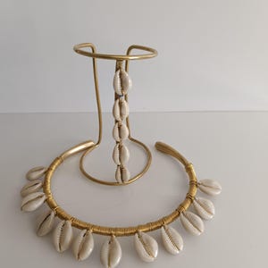 Puede incluir: Un collar dorado con conchas de cauri blancas y un soporte a juego. El collar presenta un diseño envuelto en alambre con conchas espaciadas. El soporte tiene una parte superior circular y una barra vertical con conchas.