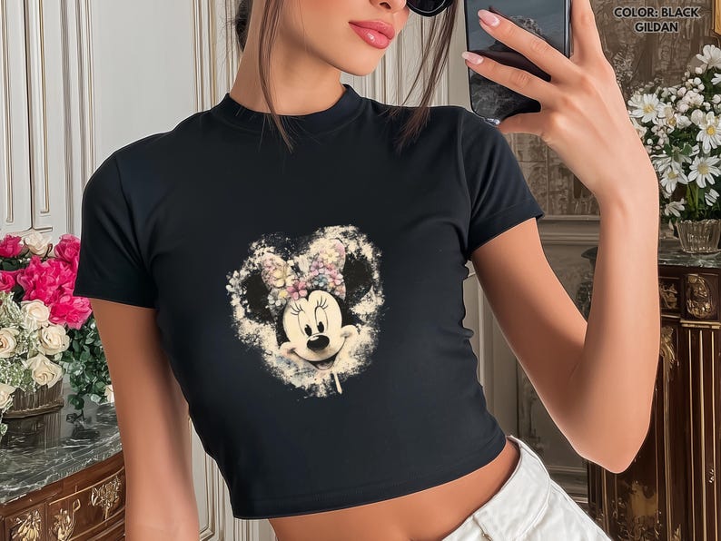 May include: Black crop top featuring a Minnie Mouse graphic. The graphic is a heart-shaped design with Minnie's face and a floral bow. The shirt has short sleeves and a mock neck. The text "COLOR: BLACK GILDAN" is in the upper right corner.