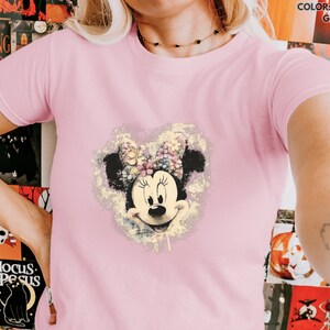 May include: Light pink t-shirt featuring a Minnie Mouse graphic. The graphic shows Minnie Mouse's face and bow, surrounded by a floral design. The text "COLOR: LIGHT PINK GILDAN" is in the upper right corner.
