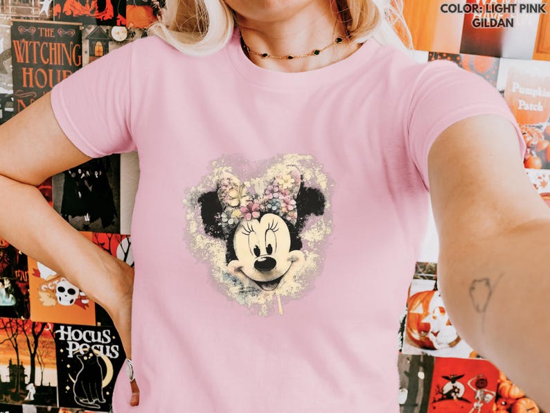May include: Light pink t-shirt featuring a Minnie Mouse graphic. The graphic shows Minnie Mouse's face and bow, surrounded by a floral design. The text "COLOR: LIGHT PINK GILDAN" is in the upper right corner.