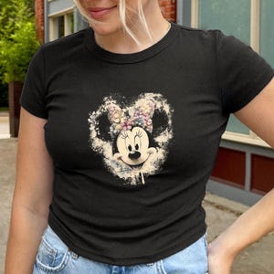 May include: Black crew neck t-shirt featuring a distressed graphic of Minnie Mouse with a floral bow. The graphic is in a heart shape. The shirt is paired with blue denim jeans.