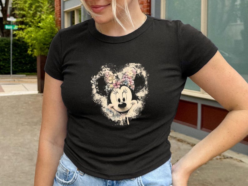 May include: Black crew neck t-shirt featuring a distressed graphic of Minnie Mouse with a floral bow. The graphic is in a heart shape. The shirt is paired with blue denim jeans.