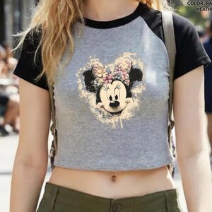 May include: A gray raglan athletic heather and black crop top featuring a Minnie Mouse graphic. The graphic is a vintage-style illustration of Minnie Mouse's face with a floral bow. The top has black sleeves and a black neckline.