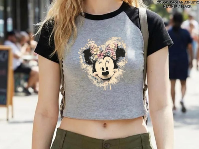 May include: A gray raglan athletic heather and black crop top featuring a Minnie Mouse graphic. The graphic is a vintage-style illustration of Minnie Mouse's face with a floral bow. The top has black sleeves and a black neckline.