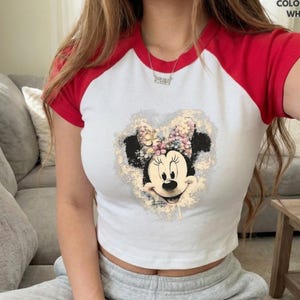 May include: White and red raglan crop top featuring a Minnie Mouse graphic. The graphic shows Minnie Mouse's face and bow with floral accents. The top has a round neckline and short sleeves.