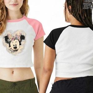 May include: White crop top with pink raglan sleeves featuring a Minnie Mouse graphic. The graphic is a heart-shaped design with Minnie's face and a floral bow. The back of the shirt is white with black raglan sleeves.
